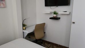 Ein kleiner, ordentlicher Raum mit einem Bett, einem Schreibtisch mit Stuhl, einem Wand-Fernseher und zwei Pflanzenregalen.