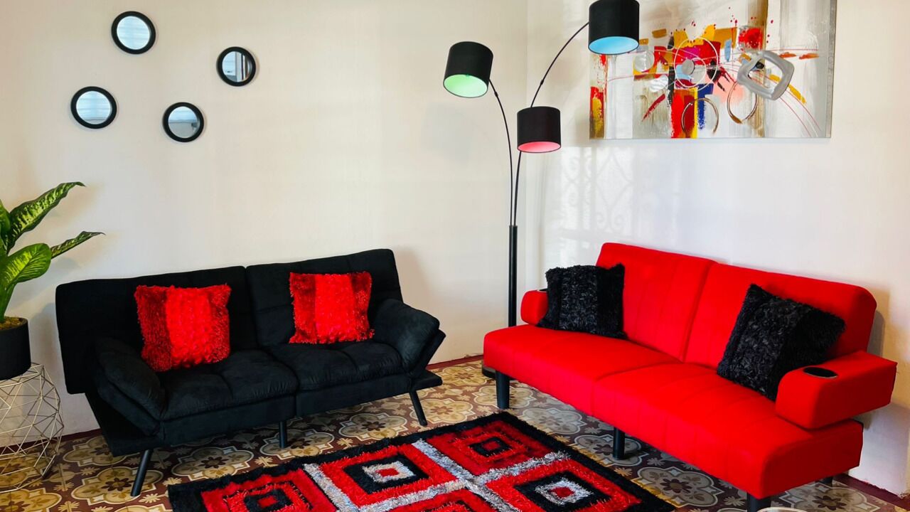 Ett vardagsrum med en svart soffa och en röd soffa, båda med röda och svarta kuddar. Det finns fyra väggspeglar och en golvlampa med flera ljusinställningar. En färgstark, abstrakt målning hänger på väggen, och en mönstrad matta ligger på golvet.