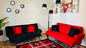 Ein Wohnzimmer mit einem schwarzen Sofa und einem roten Sofa, beide mit roten und schwarzen Kissen. Vier Wandspiegel und eine Stehlampe mit verschiedenen Lichteinstellungen sind vorhanden. Ein farbenfrohes abstraktes Gemälde hängt an der Wand, und ein gemustertes Teppich liegt auf dem Boden.