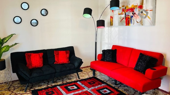 Ein Wohnzimmer mit einem schwarzen Sofa und einem roten Sofa, beide mit roten und schwarzen Kissen. Vier Wandspiegel und eine Stehlampe mit verschiedenen Lichteinstellungen sind vorhanden. Ein farbenfrohes abstraktes Gemälde hängt an der Wand, und ein gemustertes Teppich liegt auf dem Boden.