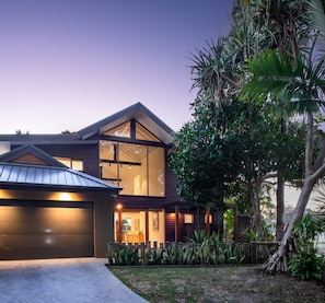 A modern two-story house with a garage, large windows, and a well-maintained garden with tropical plants. - South Straddie Retreat by Laguna Pacific (South Stradbroke)