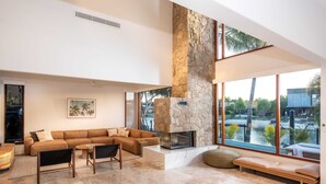 A spacious living room with a stone fireplace, large windows, and a view of a water body. - Quiet Island Paradise! (South Stradbroke)
