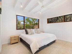 A bedroom with a bed, a nightstand, a ceiling fan, and large windows with a view of palm trees.