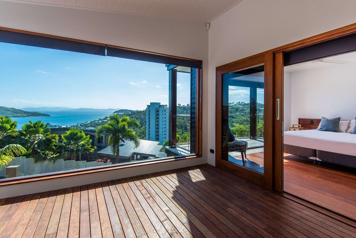 A balcony with a wooden floor and a large window offering a view of the sea and surrounding buildings.