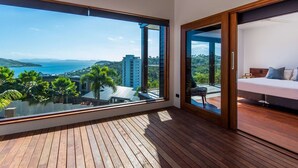 Een balkon met een houten vloer en een groot raam met uitzicht op de zee en omliggende gebouwen.