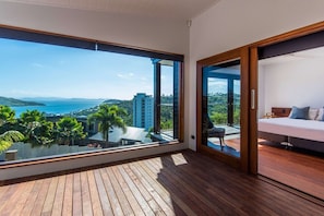 Un balcón con piso de madera y un ventanal grande que ofrece vistas al mar y edificios circundantes.