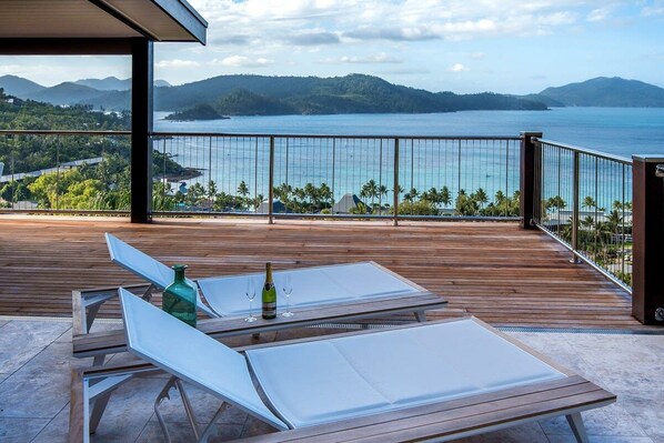 A balcony with two lounge chairs, a bottle of champagne, and a glass. The balcony overlooks a body of water and a mountainous landscape.