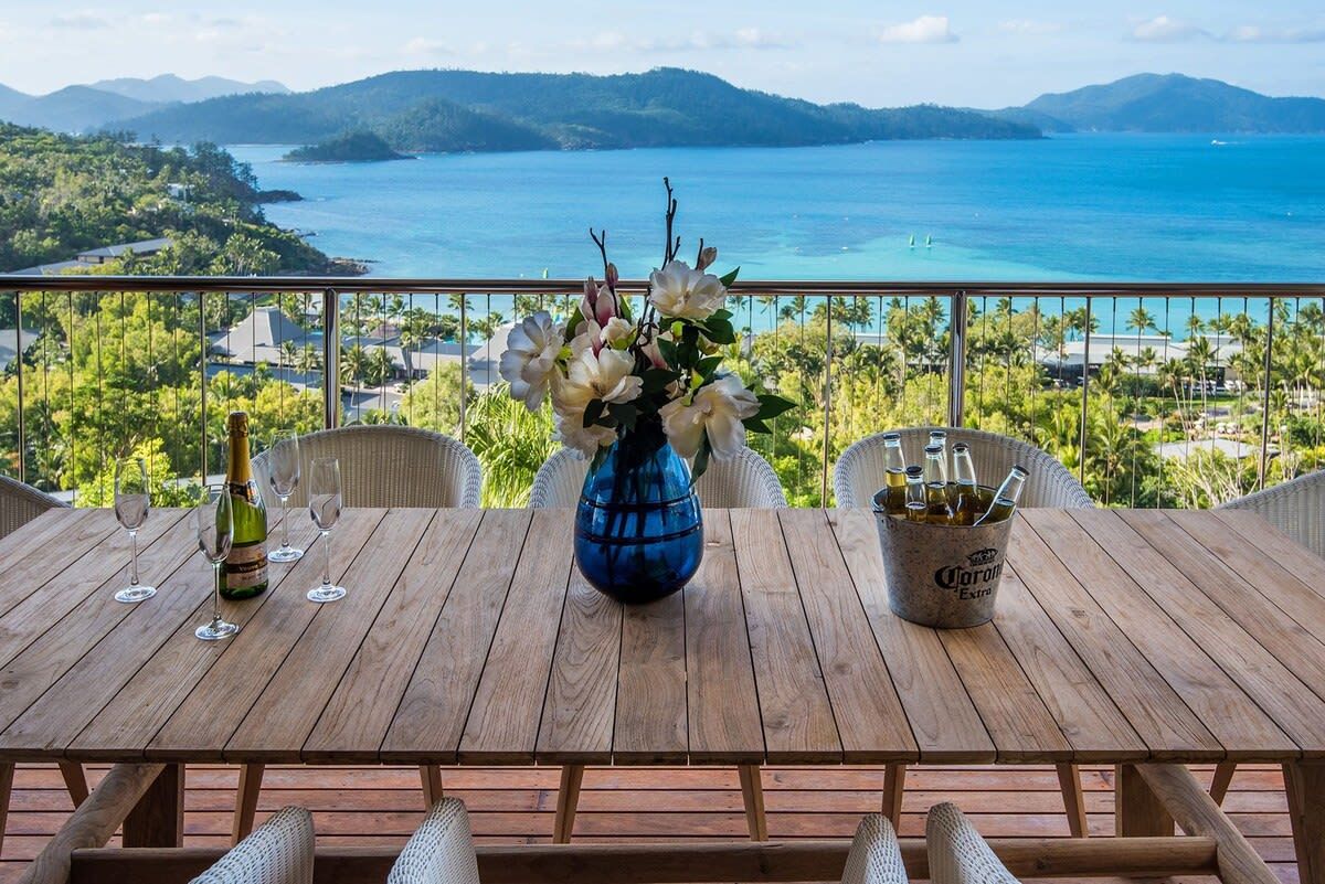 Une table en bois, sur laquelle se trouvent une bouteille de champagne, des verres et un seau de bière, offrant une vue sur la côte, avec des montagnes et une végétation luxuriante.