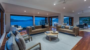 Een ruime woonkamer met een groot raam dat uitzicht biedt op zee en gebouwen, gemeubileerd met banken en fauteuils, en een salontafel.