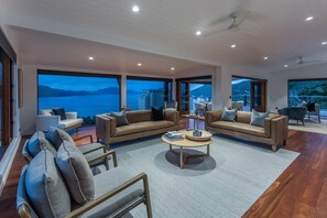A spacious living room with a large window offering a view of the sea and buildings, furnished with sofas and armchairs, and a coffee table. - Horizons - Hamilton Island (Whitsundays)