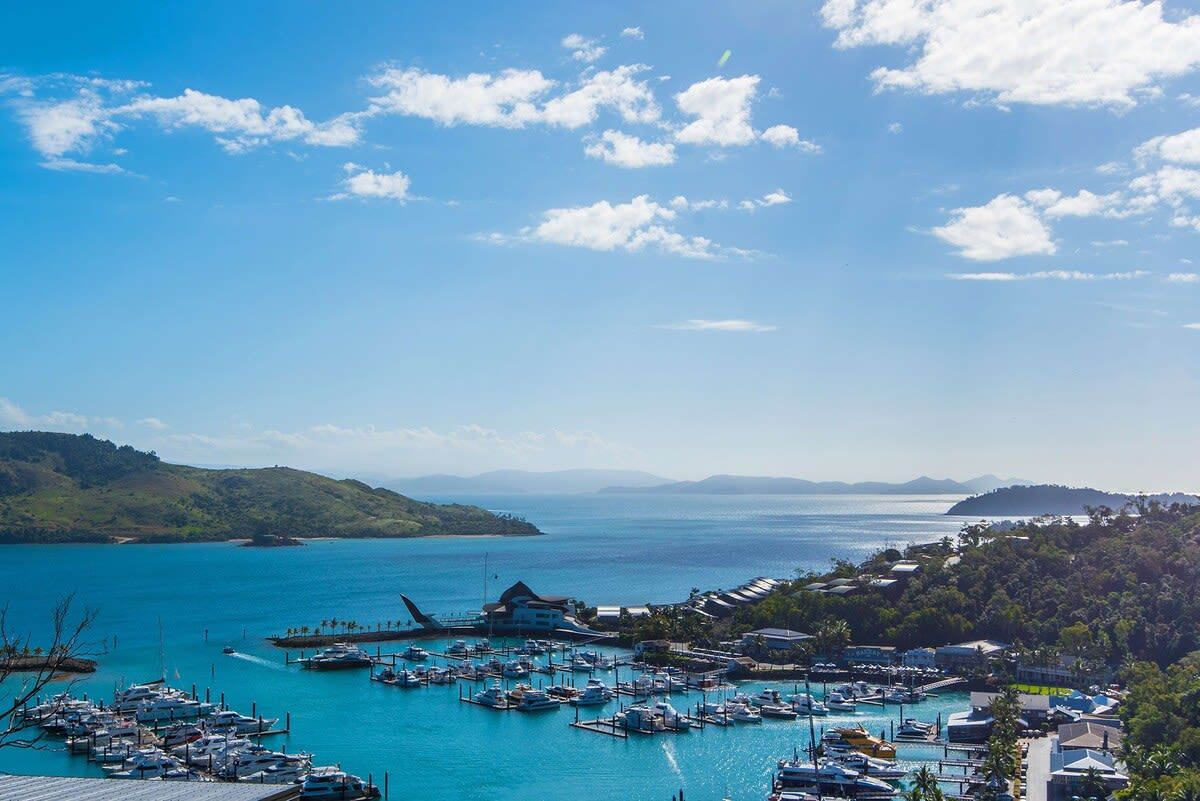 A marina with boats docked, a building with a sloped roof, and a hilly landscape in the background.