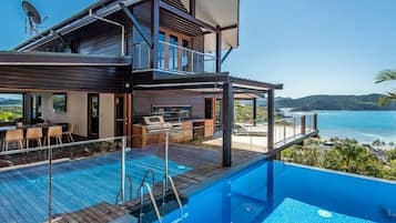 A modern two-story house with a swimming pool, outdoor seating, and a view of the ocean.
