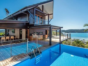 A modern two-story house with a swimming pool, outdoor seating, and a view of the ocean.
