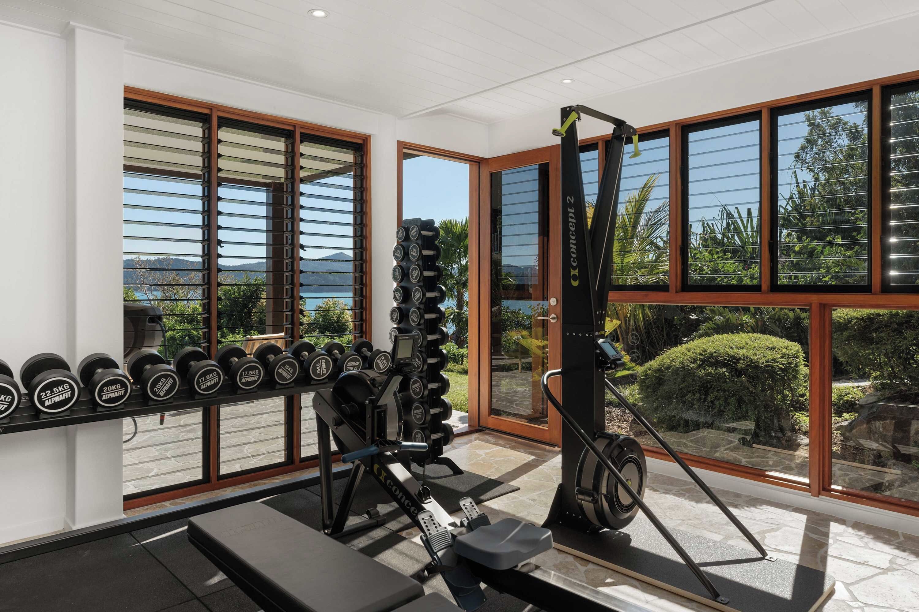 Ein Fitnessraum mit Trainingsgeräten, einer Bank und großen Fenstern mit Blick ins Freie.