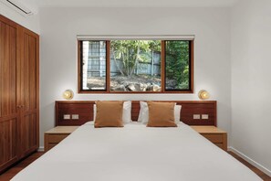 A bedroom with a large bed, wooden headboard, two bedside tables, and a window with a view of a garden. - Papillon - Hamilton Island (Whitsundays)