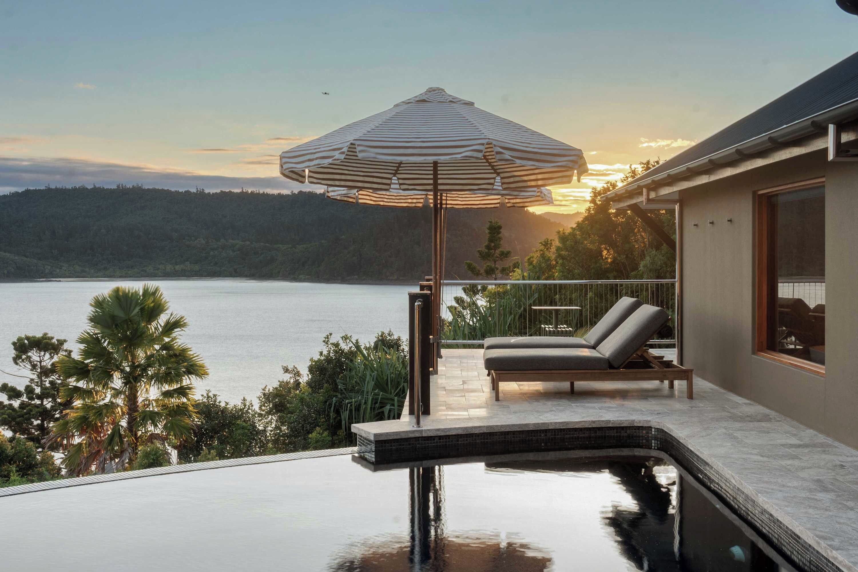 A poolside area with a view of the water, a lounge chair, and a striped umbrella.