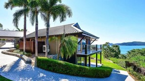 Een modern huis met een groot balkon, omgeven door weelderige begroeiing en uitzicht op zee.