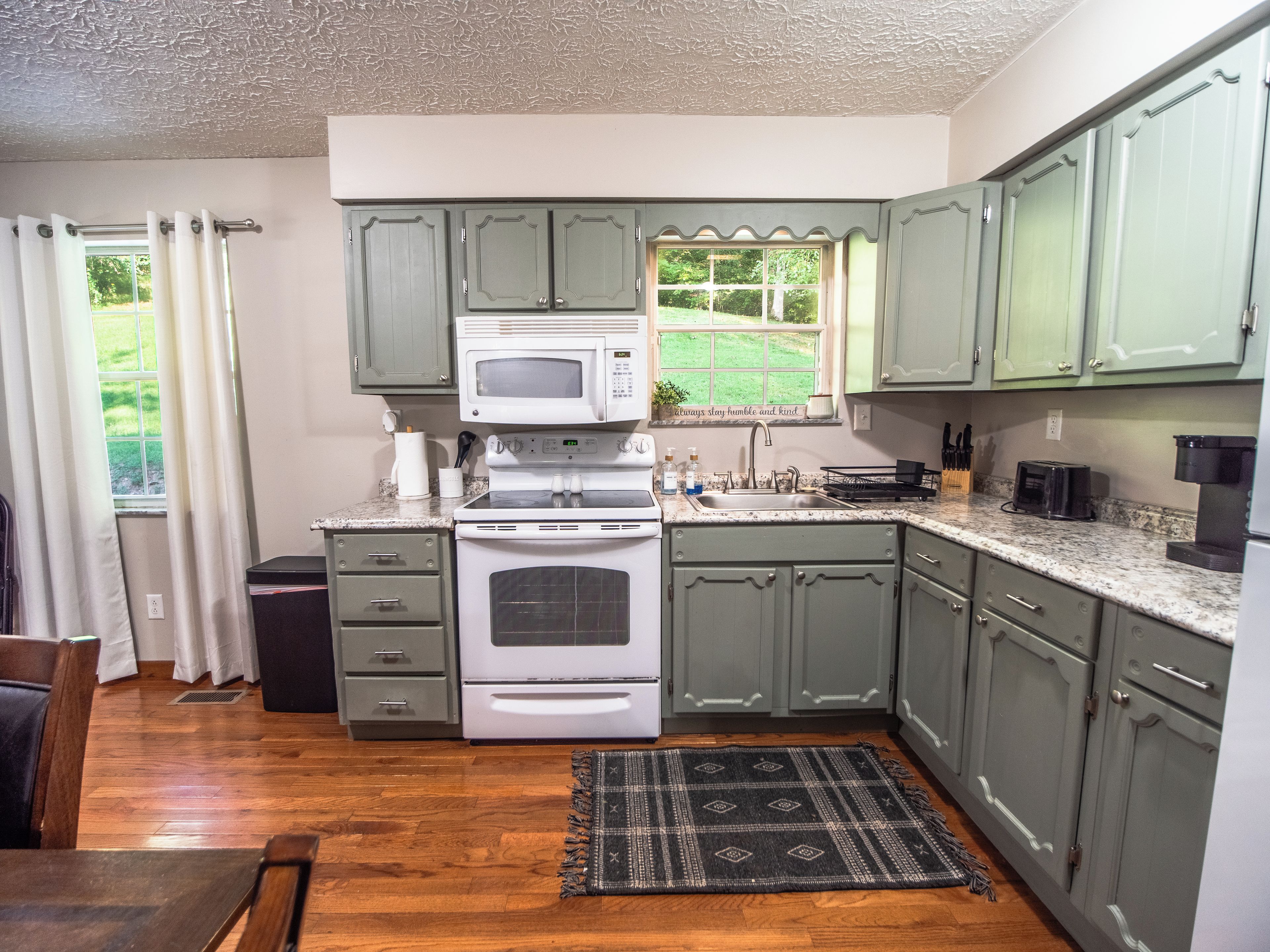 A kitchen with green cabinets, a microwave, stove, and a window with a view of greenery.
