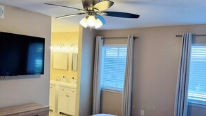 Ein Hotelzimmer mit einem Bett, einem Deckenventilator, einem Fernseher und einem Badezimmer mit Spiegel sowie Vorhängen.