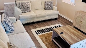 Ein Wohnzimmer mit einem beige gefärbten Sofa, gemusterten Kissen, einem Holztisch, einem Deckenventilator und einem Fenster mit Jalousien.