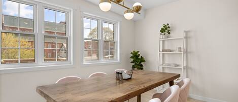 Un comedor con una mesa de madera, seis sillas, una lámpara colgante, una planta y un estante blanco.