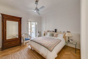 A bedroom with a large wooden wardrobe, a bed with a quilt, a chair, a glass side table, and a ceiling fan.