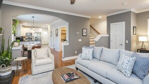 Una sala moderna con un sofá, sillones y una mesa de centro. Se ve una escalera que sube a otro nivel, y un área de cocina al fondo.