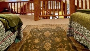 An attic bedroom with a sloped ceiling, wooden beams, a central wooden beam, a ceiling fan, a wooden railing, and two beds with patterned bedding. - Little Cottage On Prairie - Prairie Lake That Is!!! (Cameron)