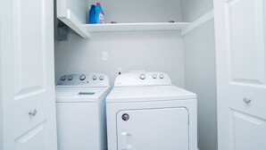 Un cuarto de lavandería con dos lavadoras y secadoras apiladas, un estante con botellas de detergente y una puerta blanca.