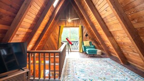 Un dormitorio acogedor en el ático con vigas de madera, un ventanal grande, un telescopio, un televisor y una silla verde.