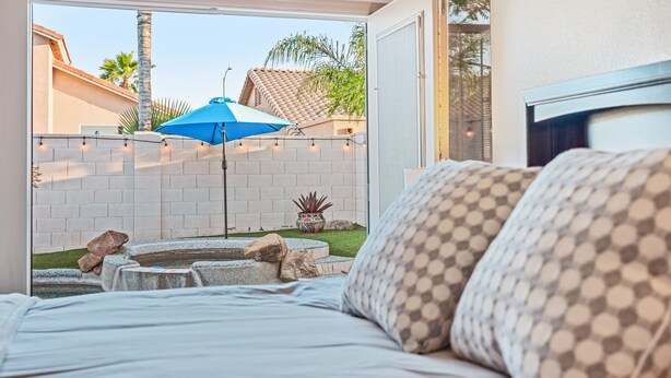 A bedroom with a view of a backyard patio, a bed with patterned pillows, and a blue umbrella. - Rose Garden Paradise (Peoria)