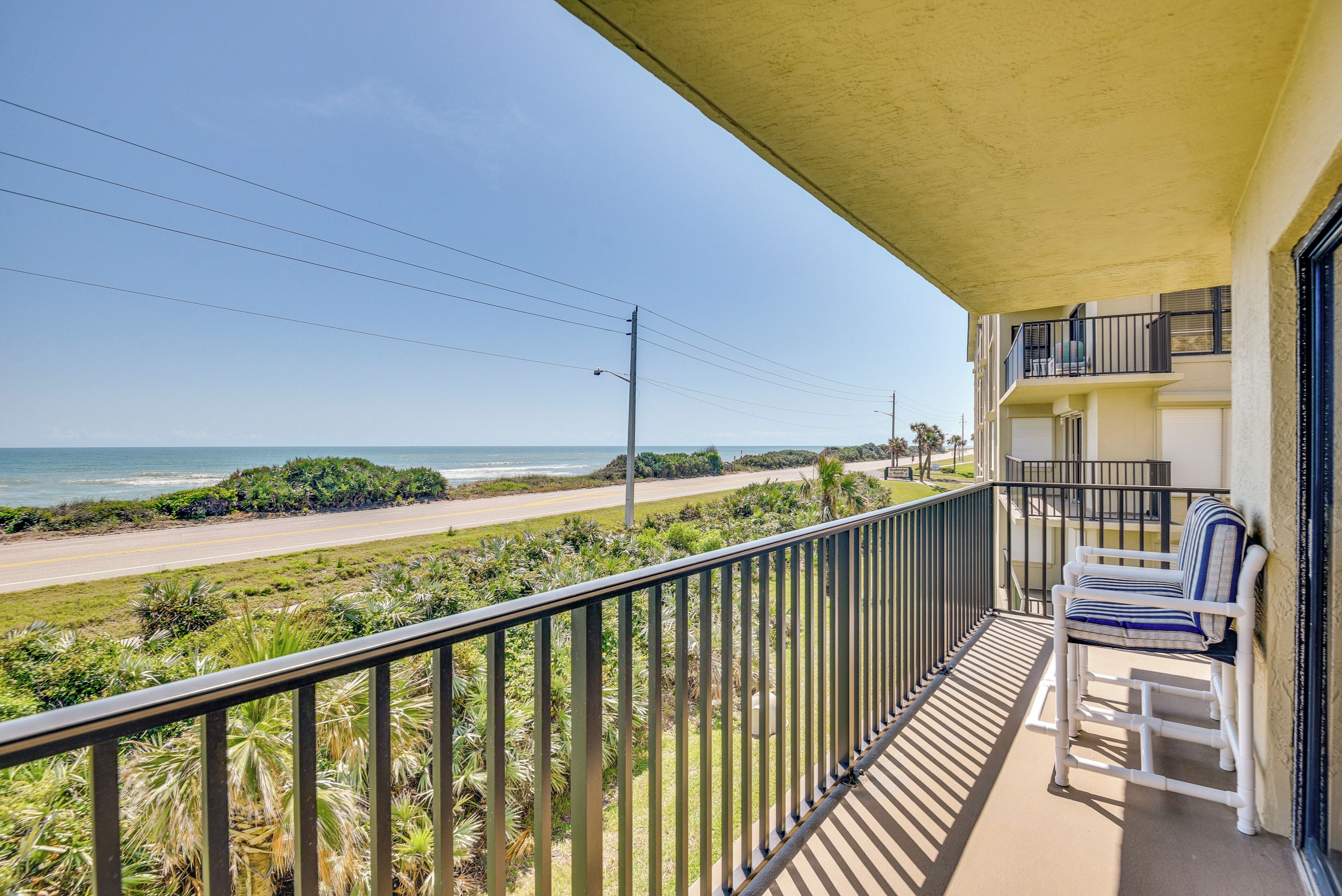A balcony with a view of the beach and ocean, a railing, and a chair.