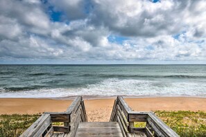 Ein hölzerner Steg führt zu einem Strand, wo Wellen an das Ufer schlagen, unter einem bewölkten Himmel.
