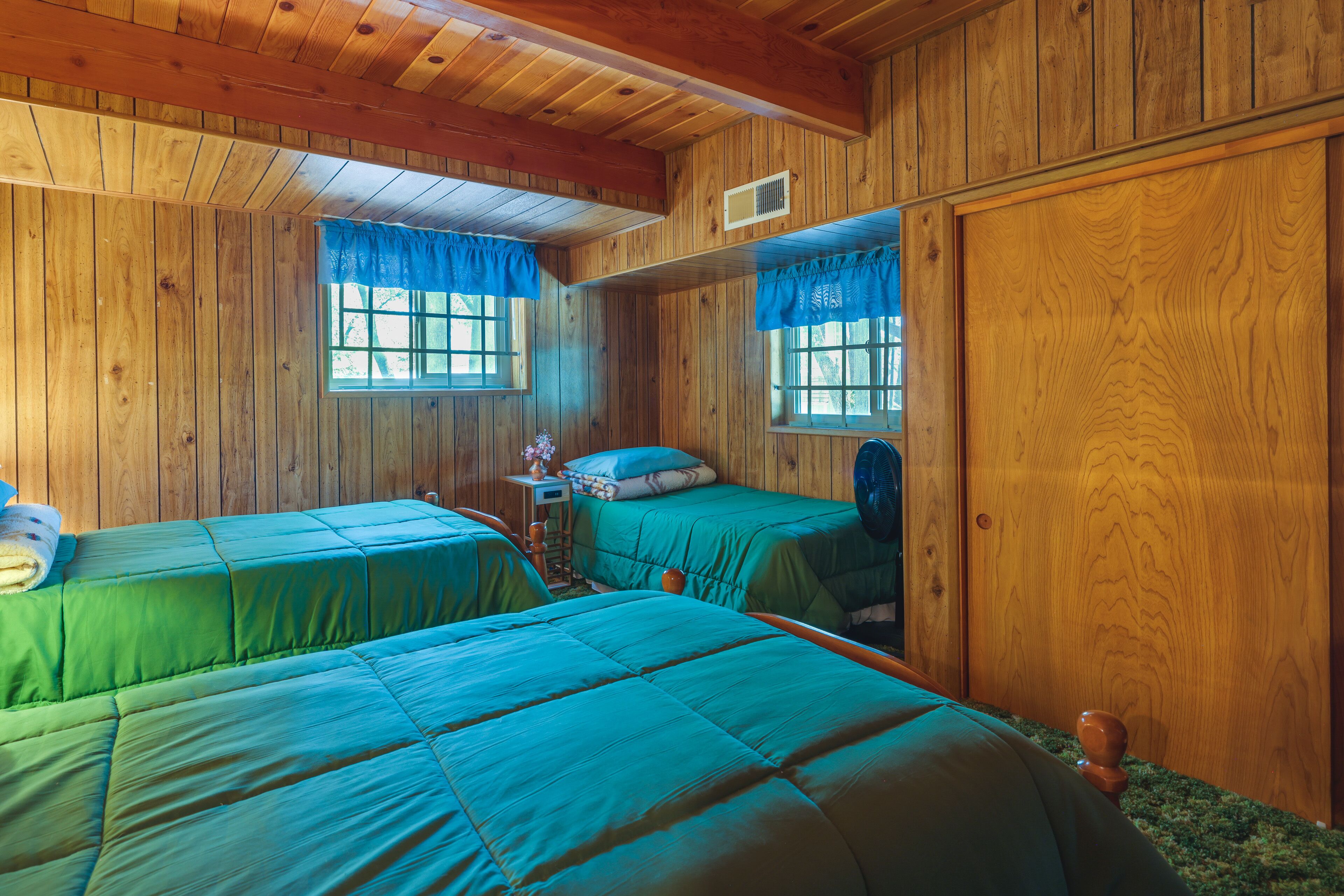 Une cabane avec deux lits, des murs en bois et une fenêtre ornée de rideaux bleus.