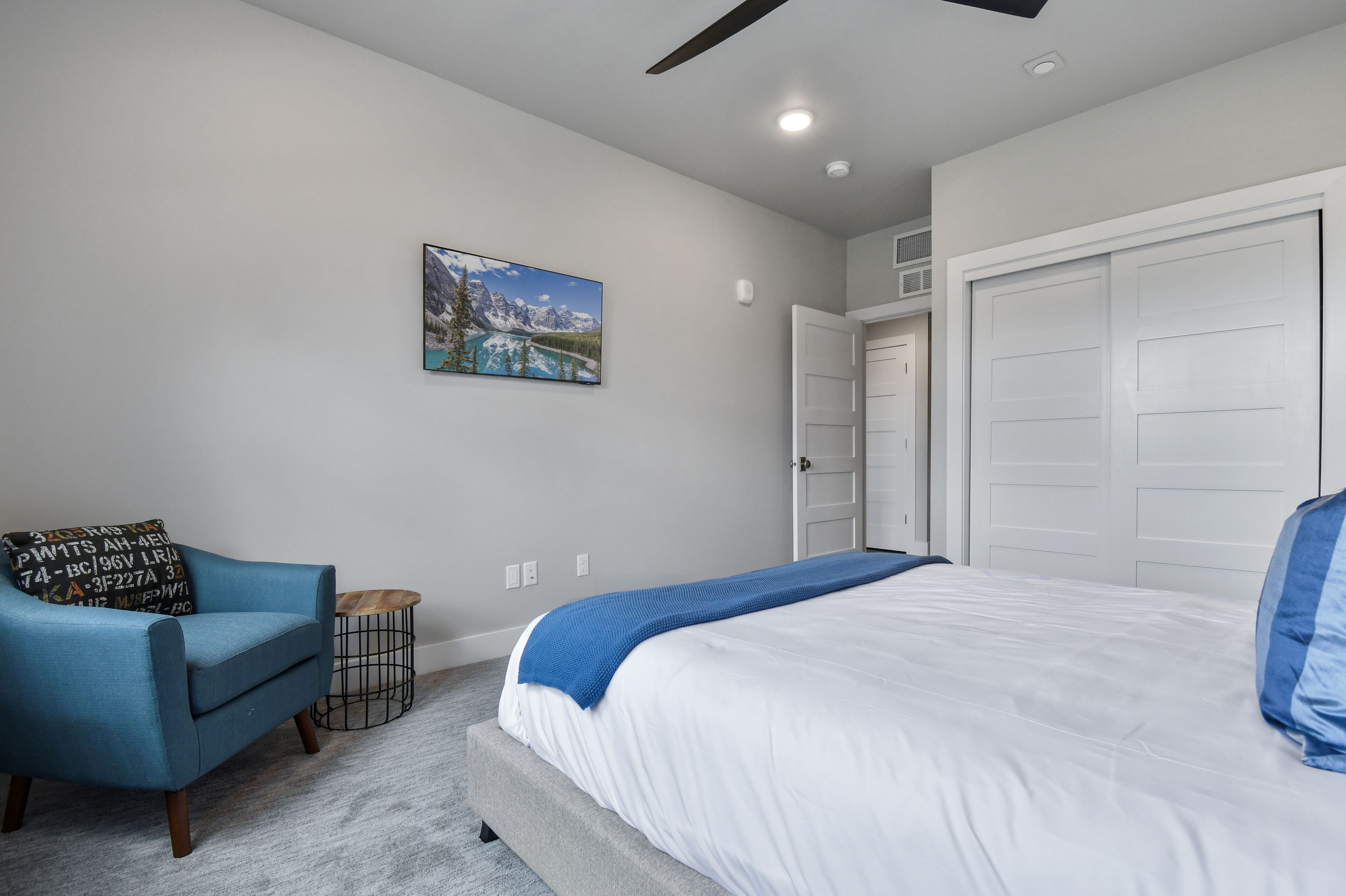 Un dormitorio con una cama, un sillón azul, una mesita auxiliar, un cuadro y una puerta blanca.