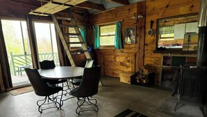 Ein Holzblockhaus-Interieur mit Esstisch, Stühlen, einer Leitern und einem Kamin, mit Blick ins Freie.
