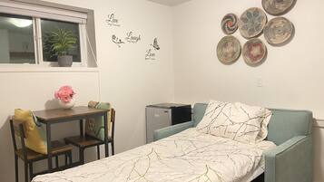 A compact room with a bed, a small table with a vase of flowers, a chair, a desk, a window with a plant, and decorative wall art.