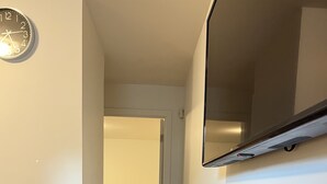 Un couloir au plancher en bois mène à une chambre à coucher où se trouvent un lit et une chaise. Une horloge est fixée au mur.