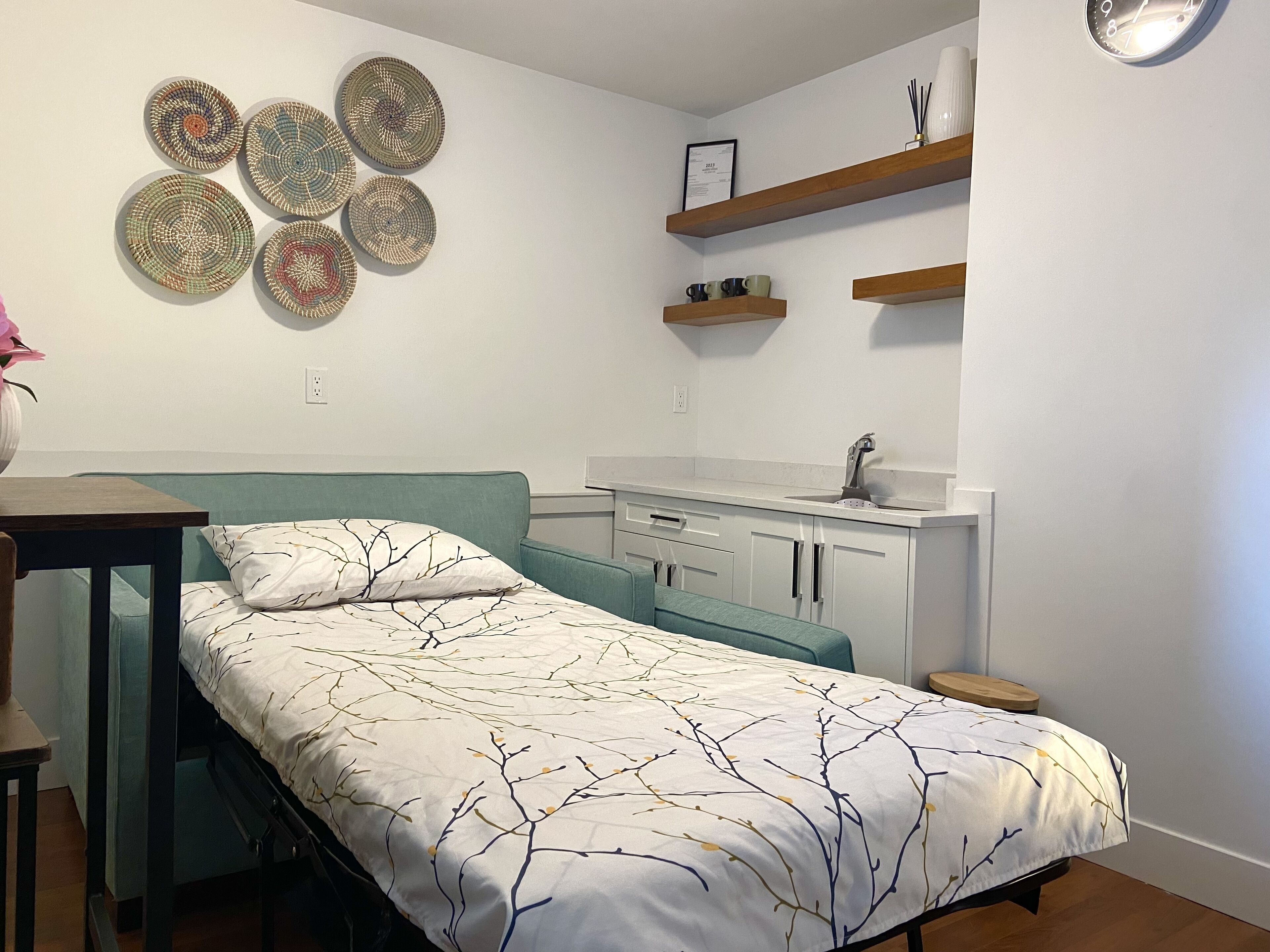 A small, neatly organized bedroom with a bed, a desk, a chair, a bedside table, a wall-mounted shelf, a wall clock, and a framed certificate.
