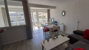 A modern living room with a white coffee table, a grey sofa with red cushions, a dining area with a white table and chairs, and a balcony with a view of buildings. - AGREABLE DEUX PIECES RENOVE DANS UNE RESIDENCE AVEC PISCINE (LE CAP D AGDE)