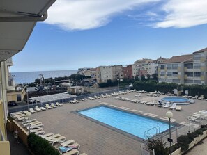 A hotel pool area with sun loungers and a swimming pool, overlooking a coastal cityscape.