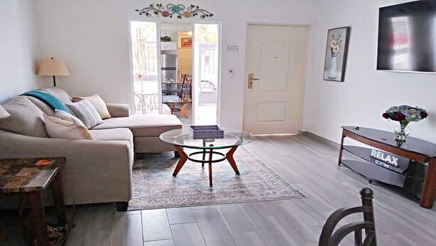 En stue med sofa, sofabord i glas, et tæppe, et tv-bord med en vase, og en dør der fører til en balkon.