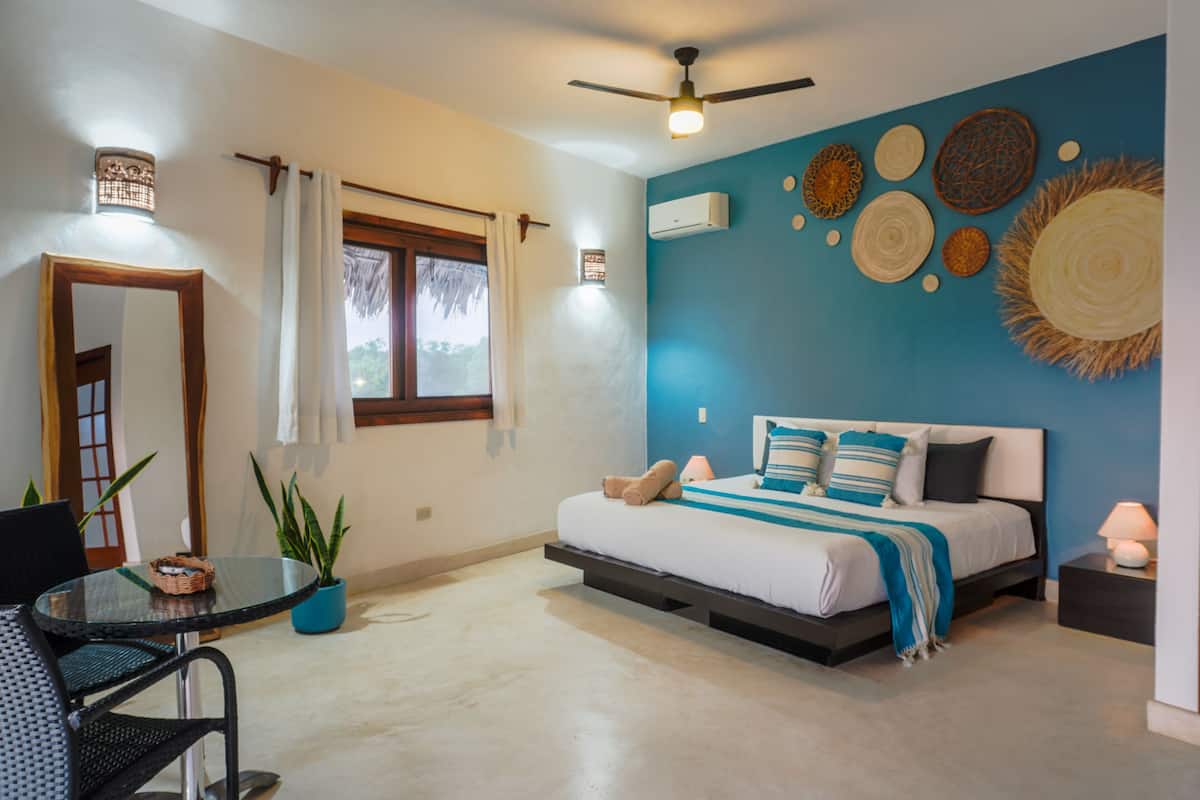 A bedroom with a bed, a ceiling fan, a window with curtains, a table with a chair, and a potted plant.