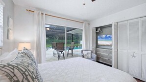 A bedroom with a bed, a TV, a wardrobe, and a sliding glass door leading to a patio with chairs and a pool.