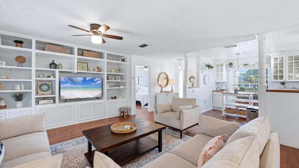 A living room with a TV, white shelves, a ceiling fan, and a view to the outside.
