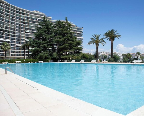 A swimming pool with lounge chairs, surrounded by palm trees and a large building with balconies. - Elegant Studio With Marina Sea And Esterel View (Mandelieu-la-Napoule)