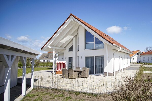 A modern two-story house with a sloped roof, large windows, and a covered patio area with outdoor furniture.