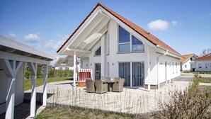 A modern two-story house with a sloped roof, large windows, and a covered patio area with outdoor furniture.
