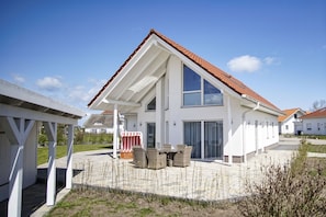 Ein modernes, zweistöckiges Haus mit Satteldach, großen Fenstern und einer überdachten Terrasse mit Gartenmöbeln.