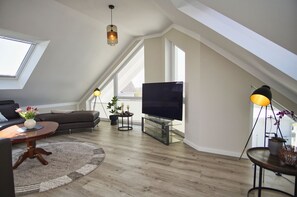 Ein modernes Wohnzimmer mit schrägverdeckter Decke, einem Flachbildfernseher, einem Sofa, einem Couchtisch und einer Stehlampe.